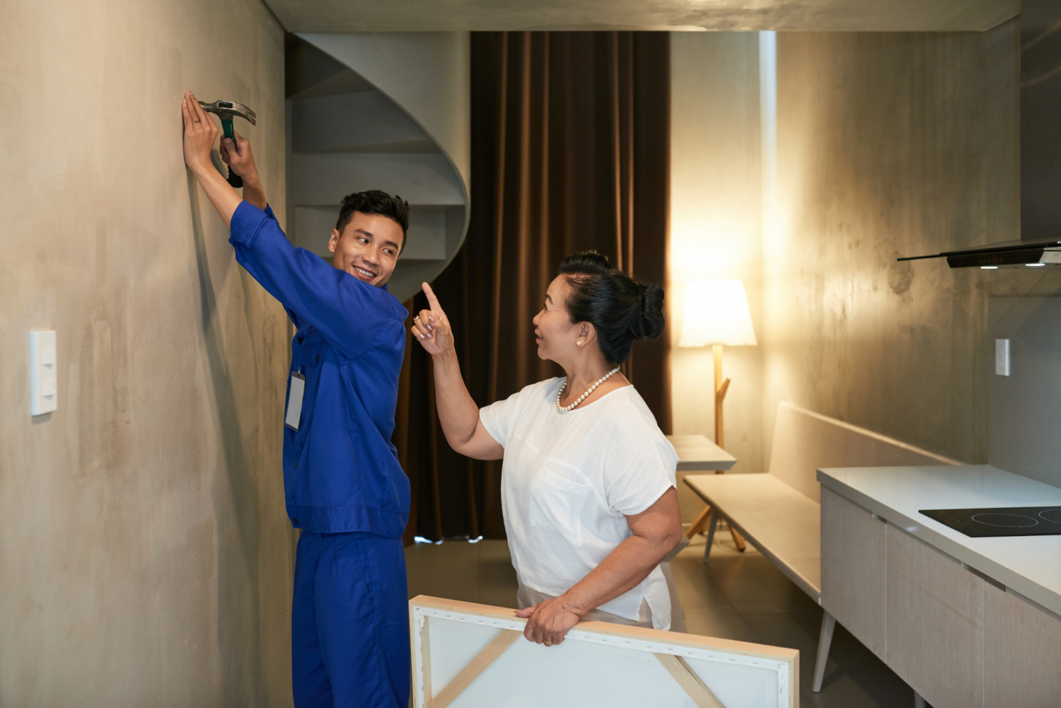 Person in blue overalls with a hammer helping set up decor in a modern living room with a woman in white supervising